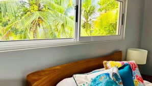 A bed with a wooden headboard, a window with a view of palm trees, and a colorful pillow. - Jaco beach apartment (Jacó)