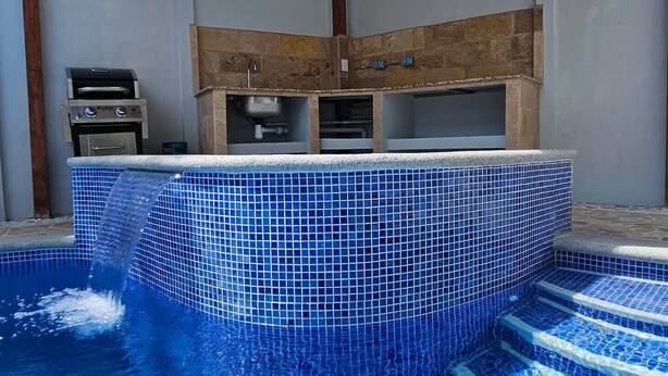 Ein Swimmingpool mit blauen Mosaikfliesen, ein Grill und eine Pergola.