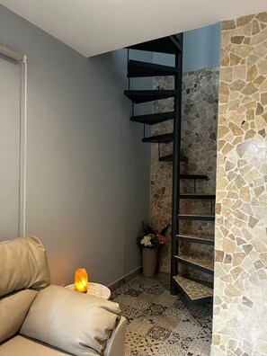 Eine Ecke eines Raumes mit einer grauen Wand, einer schwarzen Wendeltreppe, einem beigen Sofa und einem kleinen Tisch mit einer Kerze.
