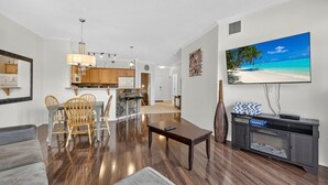 A modern living room with a sofa, dining table, and chairs, a kitchen area with wooden cabinets, and a large TV on the wall. - Downtown Pool View 2 Bdrm 449 On The Lake (Kelowna)