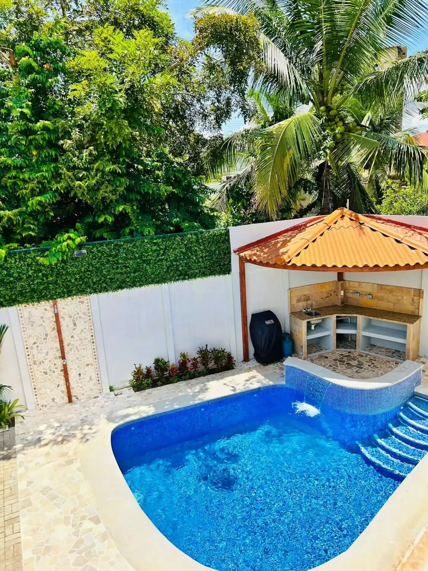 Ein Schwimmbecken mit gefliestem Deck, einer kleinen überdachten Sitzgelegenheit und üppigem Grün im Hintergrund.