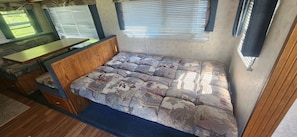 A compact RV interior with a bed, a small table, and a window with blinds.
