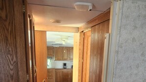 Quarto com cama, guarda-roupa de madeira e vista para uma cozinha com armários e ventoinha no teto.