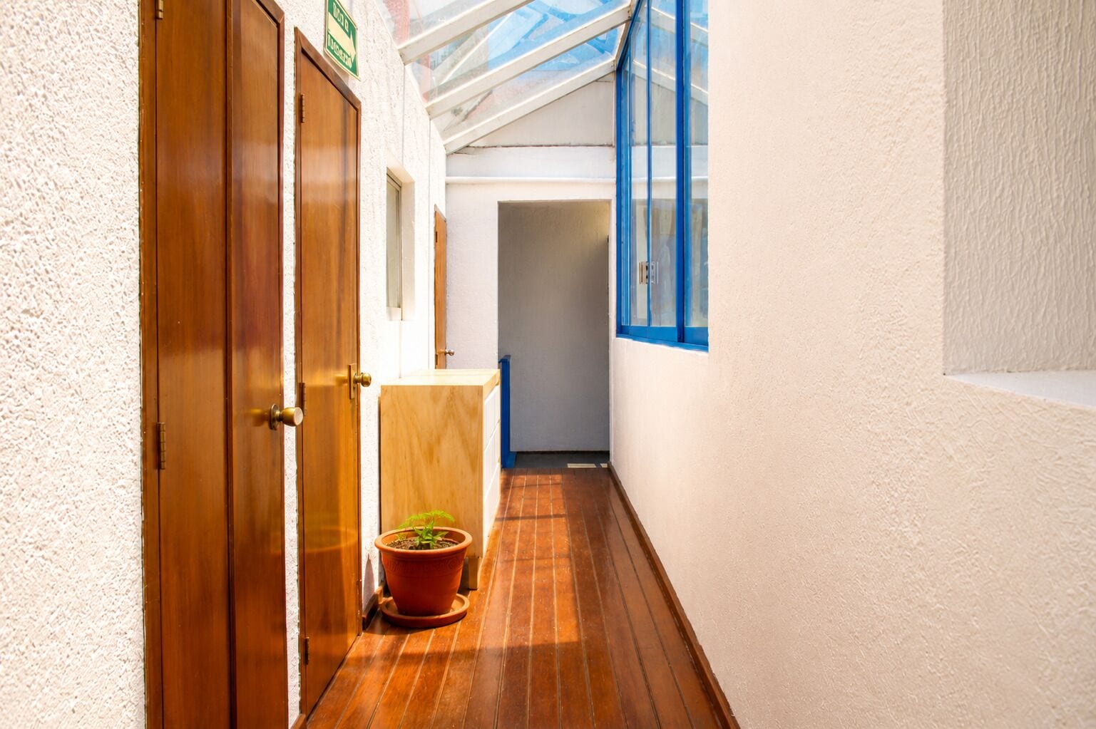 Um corredor estreito com piso de madeira, um vaso com planta e uma porta com um letreiro.