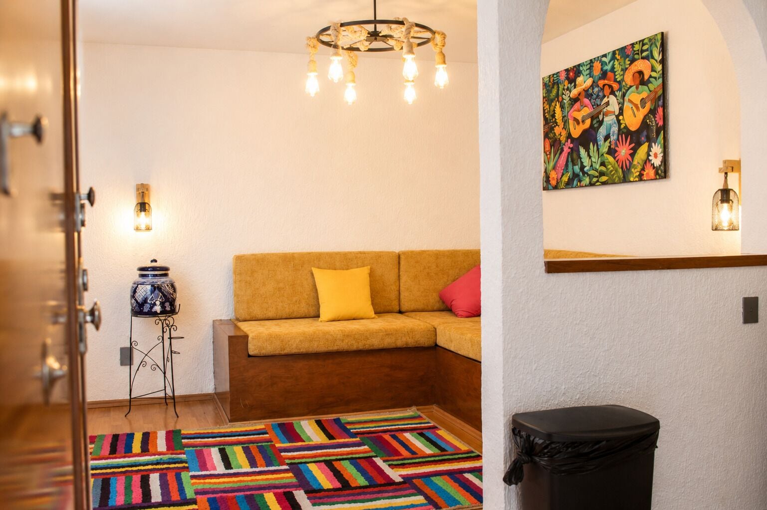 Une pièce avec un canapé, un tapis rayé aux couleurs vives, un tableau, un lustre et une poubelle.