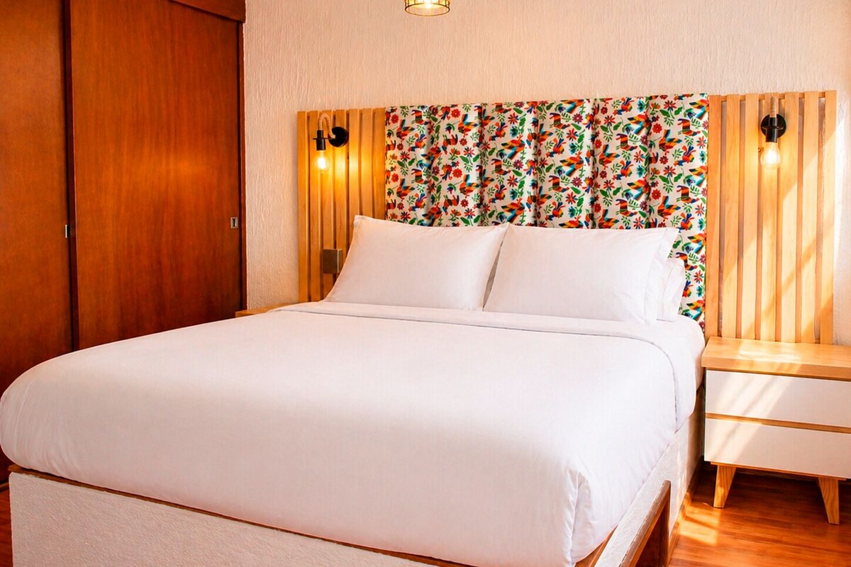 Een net opgemaakt bed met witte lakens, een houten hoofdbord met een bloemendecor en twee bedlampjes.