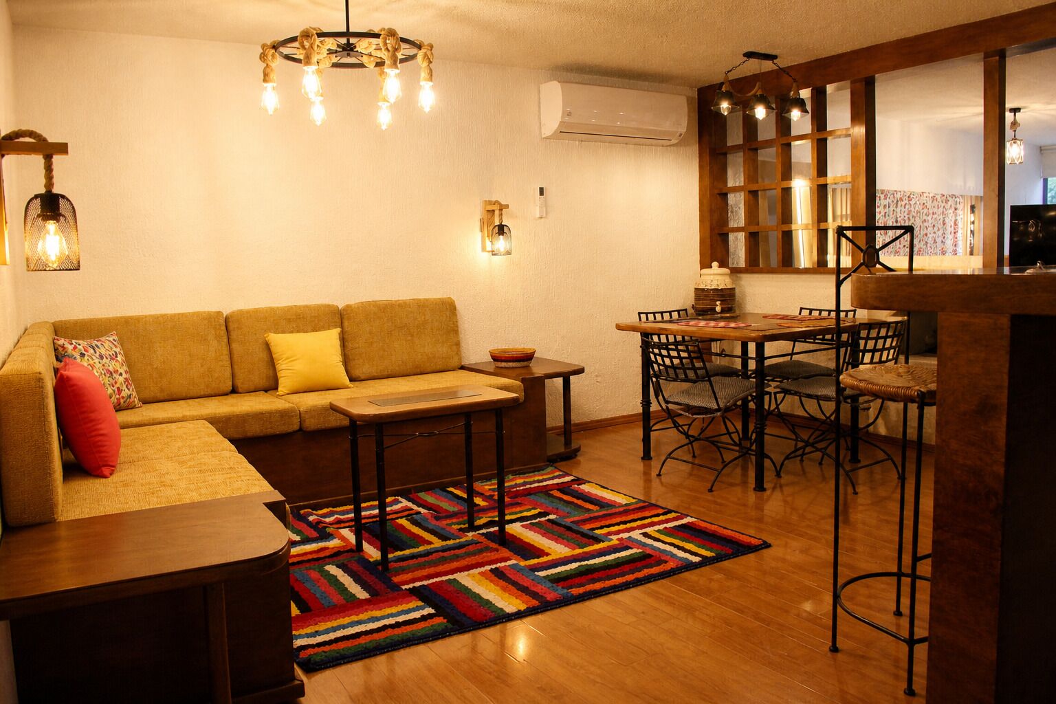 A living room with a sofa, a coffee table, a dining table with chairs, a colorful rug, and a chandelier.