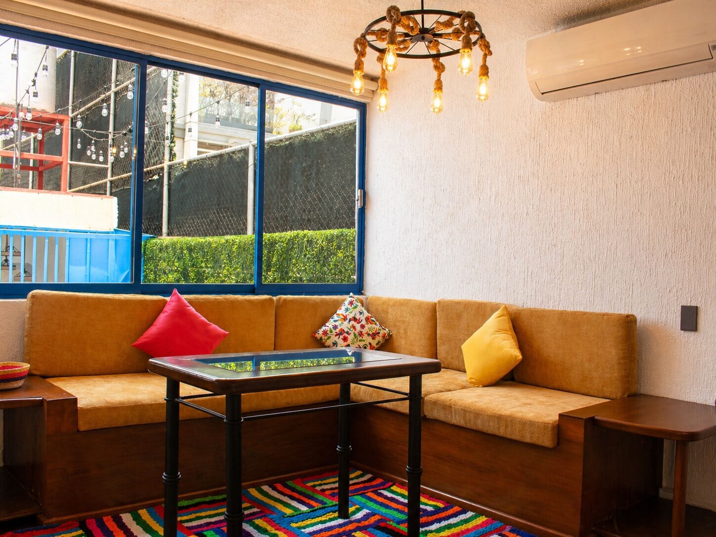 Een moderne woonkamer met een bank, een salontafel, een kleurrijk vloerkleed, een kroonluchter en een groot raam met uitzicht op een blauw gebouw.