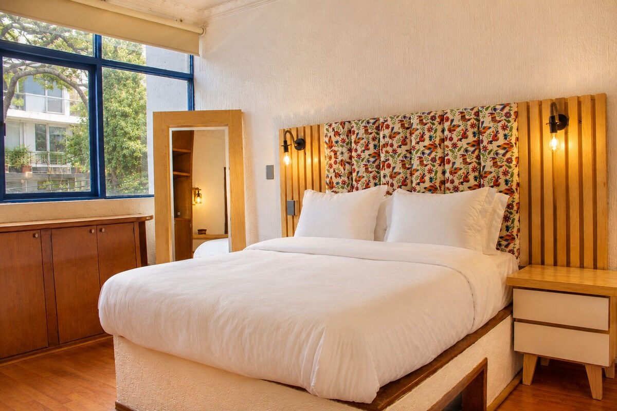Een slaapkamer met een groot bed, een houten hoofdbord, nachtkastjes en een raam met uitzicht op groen.