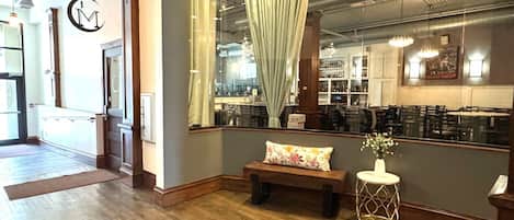 A hotel lobby with a wooden floor, a bench, a small table with a vase, and a view of the bar area through a window with curtains.