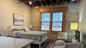 A bedroom with a bed, a chair, a desk, a window, and a brick wall. - Historic Warehouse converted into Cozy Boutique Loft 305 (Fargo)