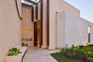 A modern building with a concrete facade, a glass door entrance, and a small garden area with plants. - Kaia House · Cap Cana Private Villa · Sleeps 14 (Punta Cana)
