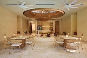 A spacious living area with a central seating arrangement, a kitchen in the background, and ceiling fans.
