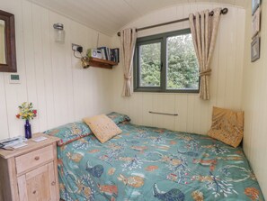 A room with a bed, a wooden dresser, a window with curtains, a shelf with books and a radio, and a wall clock. - The Three Spaniels Shepherds Hut (Llanybydder)
