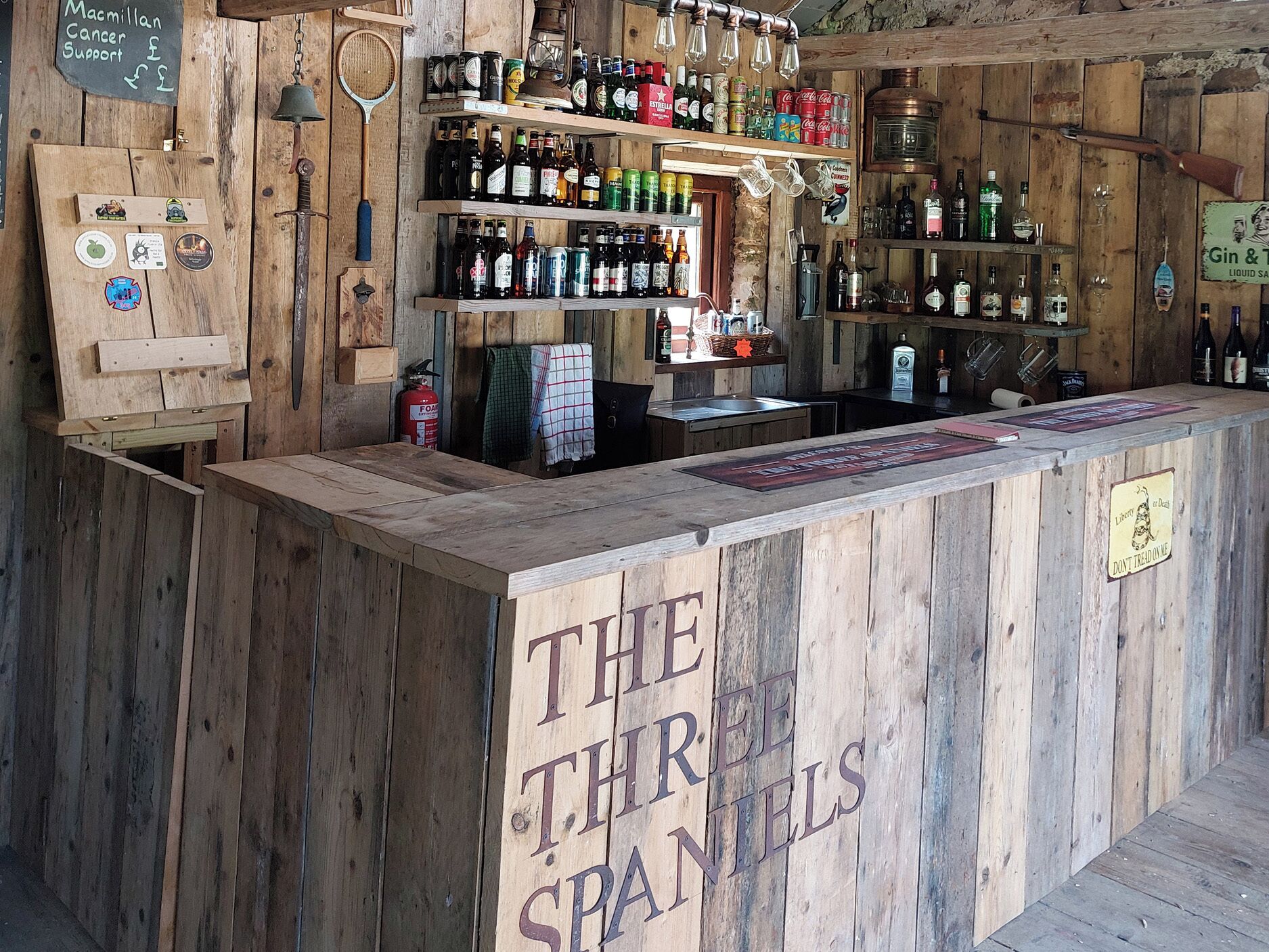 A rustic bar with wooden shelves stocked with various bottles and a sign reading 'THE THIRD SPANELS'.