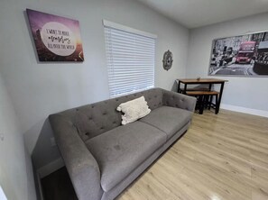 Ein Wohnzimmer mit grauem Sofa, einem kleinen Tisch, einer Wand mit Stadt-Plakat und einem Fenster mit Jalousien.
