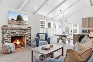 A cozy living room with a stone fireplace, a comfortable chair, a coffee table, and a view of the beach through a large window. - Coastal cottage with hot tub, close walk to beach (Cannon Beach)