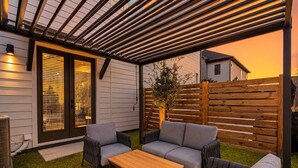 Een overdekt terras met een houten tafel en twee grijze banken, een houten hek en een huis op de achtergrond.