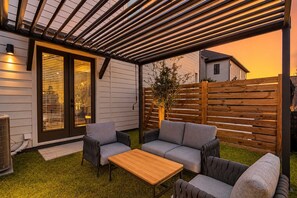 Eine überdachte Terrasse mit einem Holztisch und zwei grauen Sofas, einem Holzzaun und einem Haus im Hintergrund.