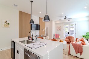A modern kitchen with a central island, a dining area with orange chairs, and a living room with a sofa and a large window. - Spring Branch 3BR - Mins to CityCentre (Houston)