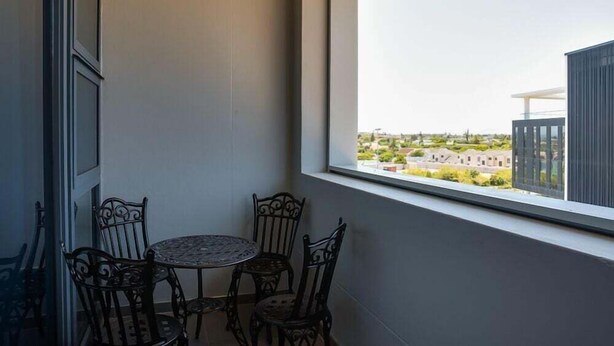 Un balcone con tavolo e sedie, una finestra che offre vista su edifici e una porta a sinistra.