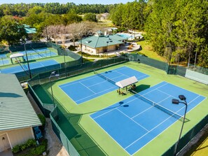 A tennis court complex with multiple courts, a covered seating area, and surrounding greenery.