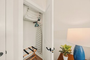 A room with a bed, a chair, a hanging laundry rack, a potted plant, and a lamp.