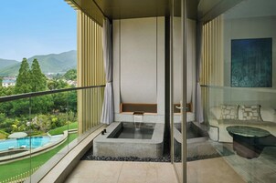 Un balcón moderno con jacuzzi, barandilla de vidrio y vistas a una piscina y follaje.
