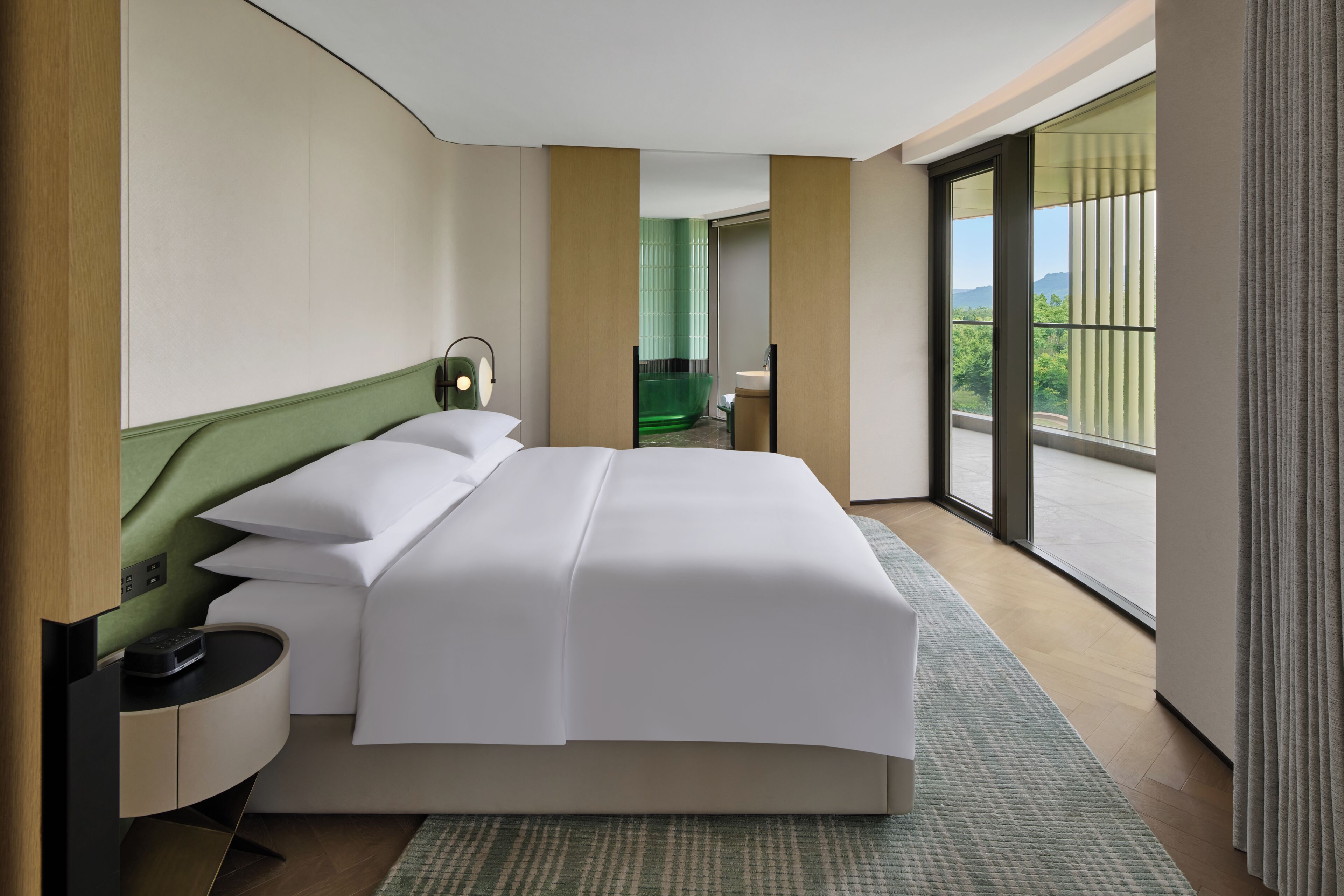 A modern hotel room with a large bed, a bedside table, a lamp, and a view of greenery through the window.