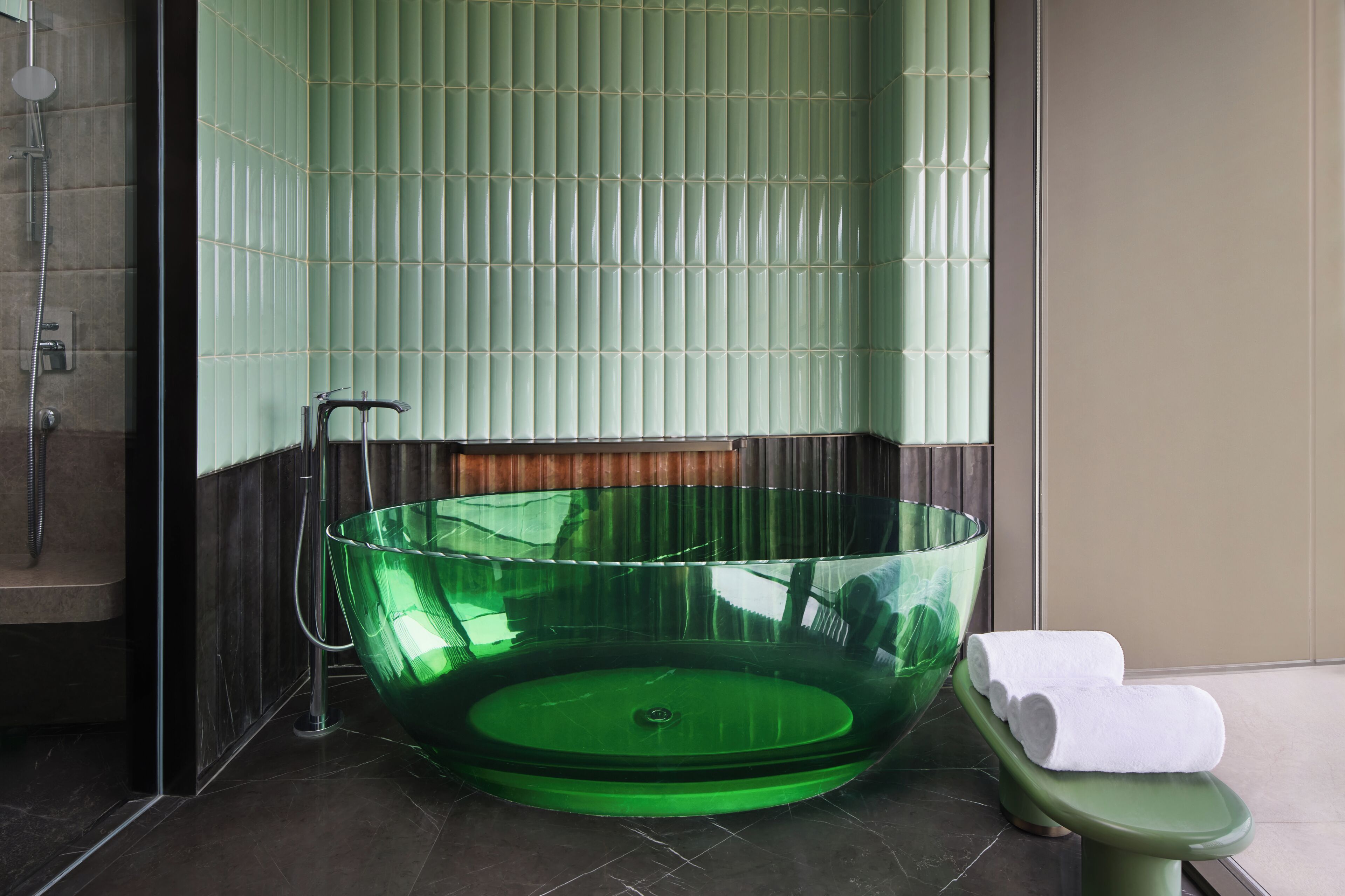 Un baño moderno con una bañera de vidrio verde, una ducha y una silla verde con una toalla blanca.