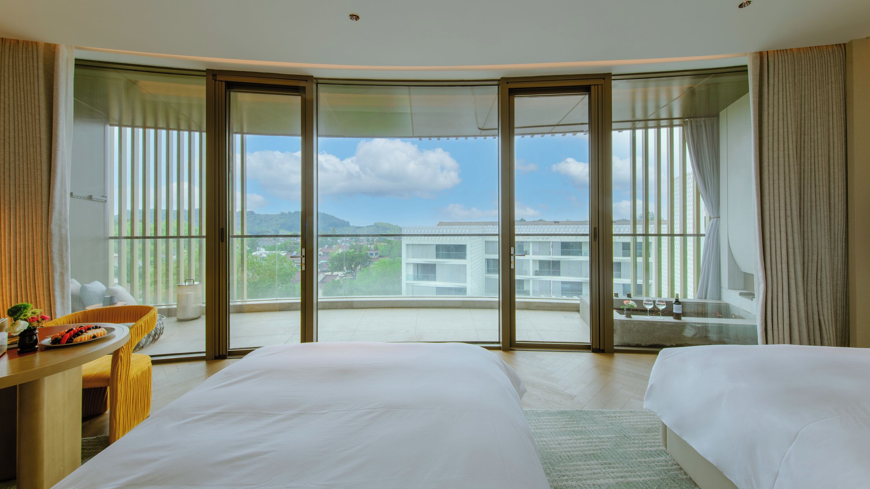 Ein Hotelzimmer mit zwei Betten, einem Schreibtisch und einem Balkon mit Blick auf Gebäude und Grünflächen.