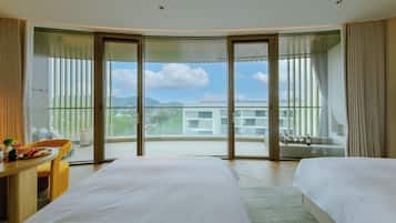 Ein Hotelzimmer mit zwei Betten, einem Schreibtisch und einem Balkon mit Blick auf Gebäude und Grünflächen.