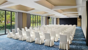 Un salón de conferencias con filas de sillas blancas, ventanas amplias y una alfombra azul.