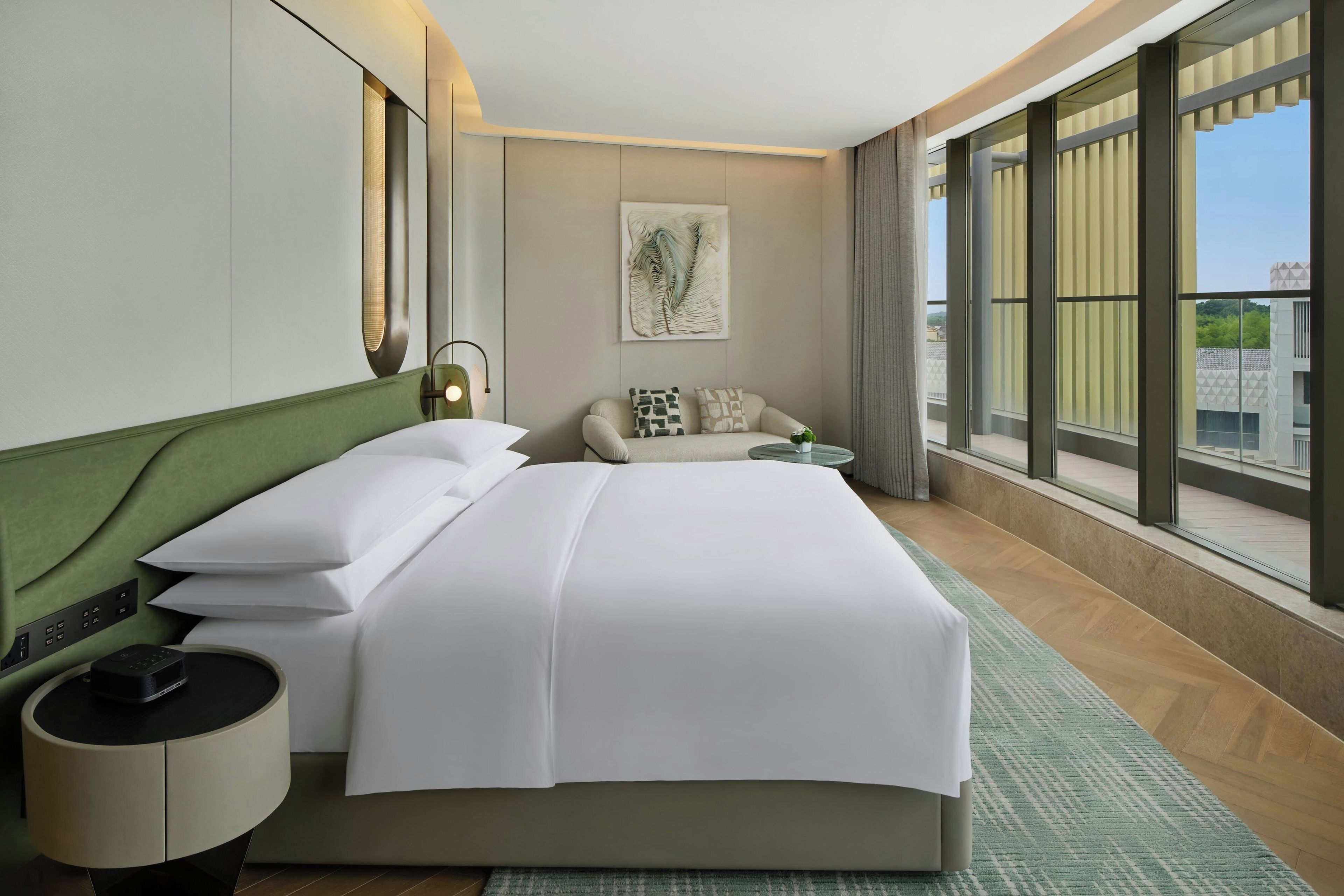 A hotel room with a large bed, a bedside table, a lamp, a sofa, and a large window offering a view.