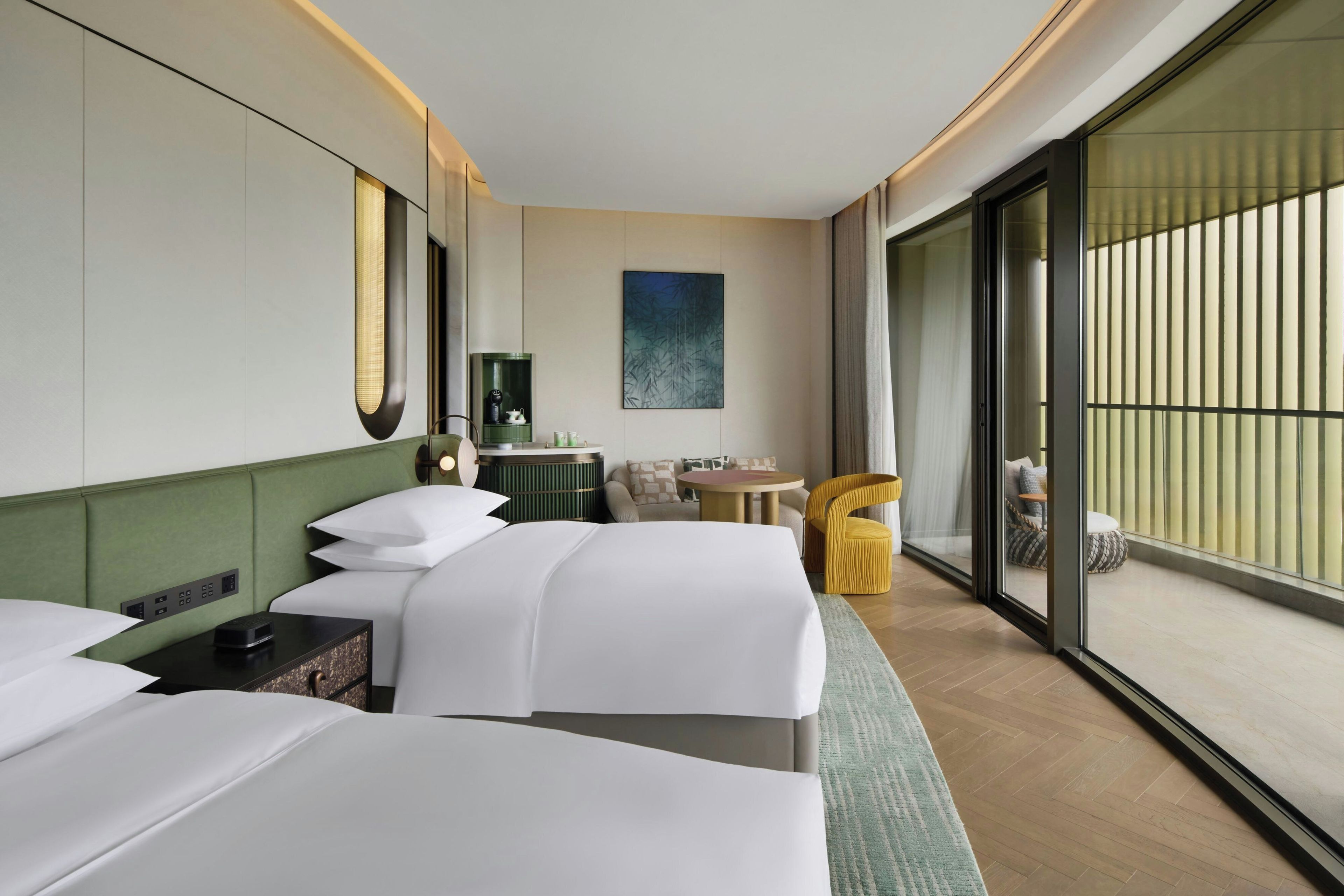 Habitación de hotel con dos camas, una mesita, una silla, un sofá, un cuadro y un ventanal amplio.