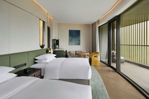 Habitación de hotel con dos camas, una mesita, una silla, un sofá, un cuadro y un ventanal amplio.