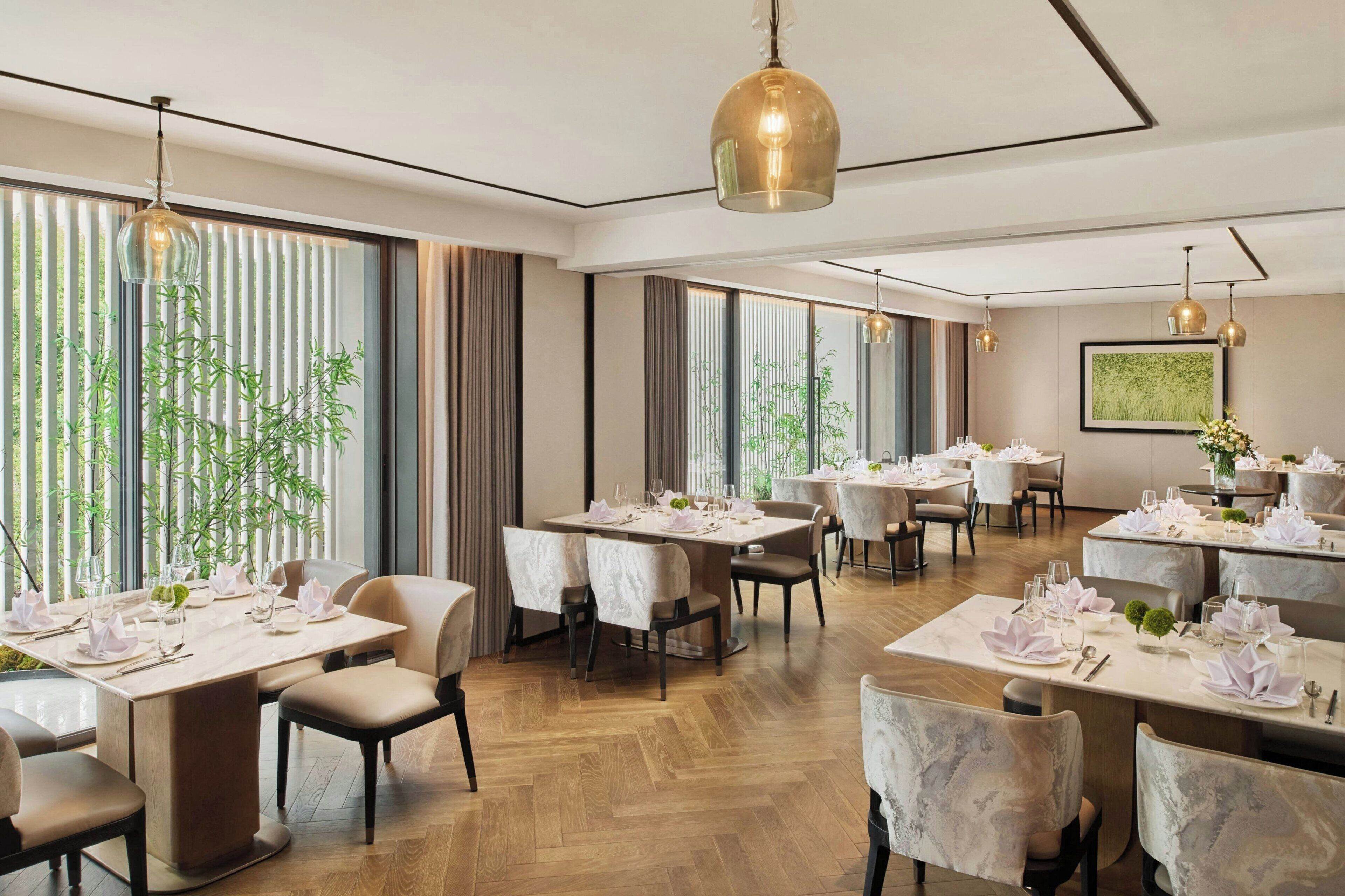 Un restaurante con varias mesas dispuestas para comer, amplias ventanas con persianas verticales y lámparas colgantes.
