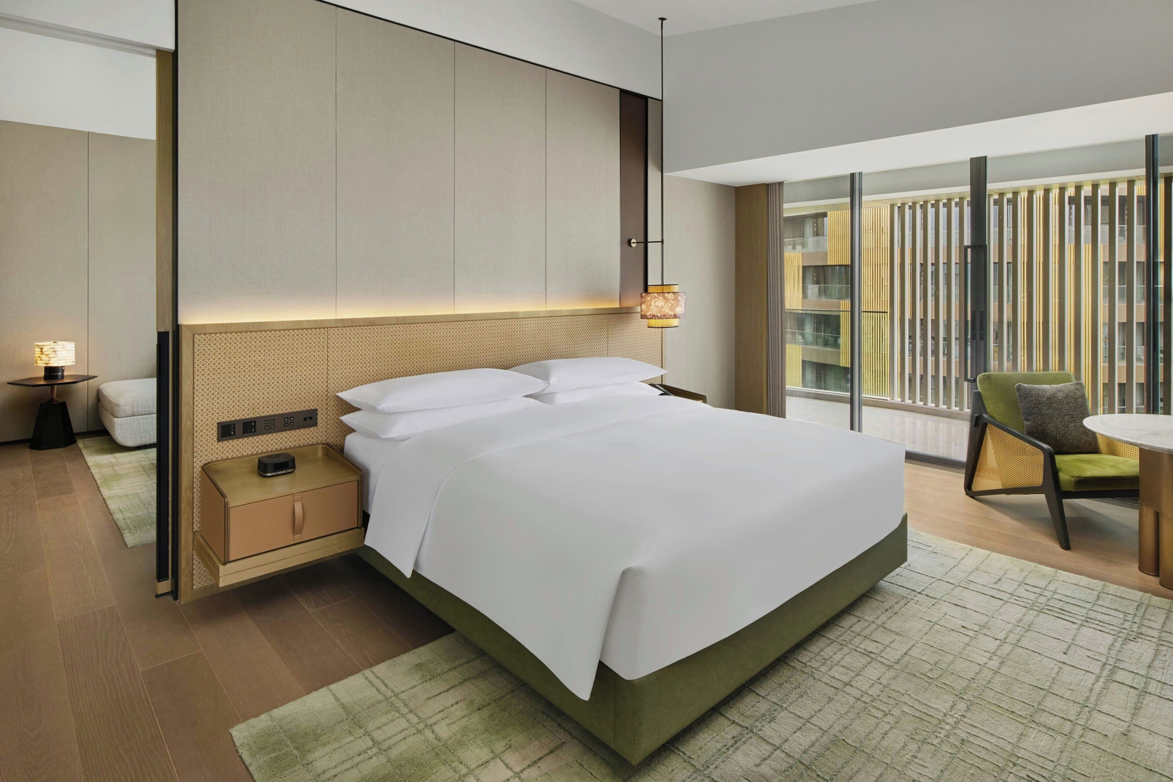 Una habitación de hotel moderna con una cama grande, mesitas de noche, una lámpara, una pequeña mesa con una silla y vistas a un edificio a través de la ventana.