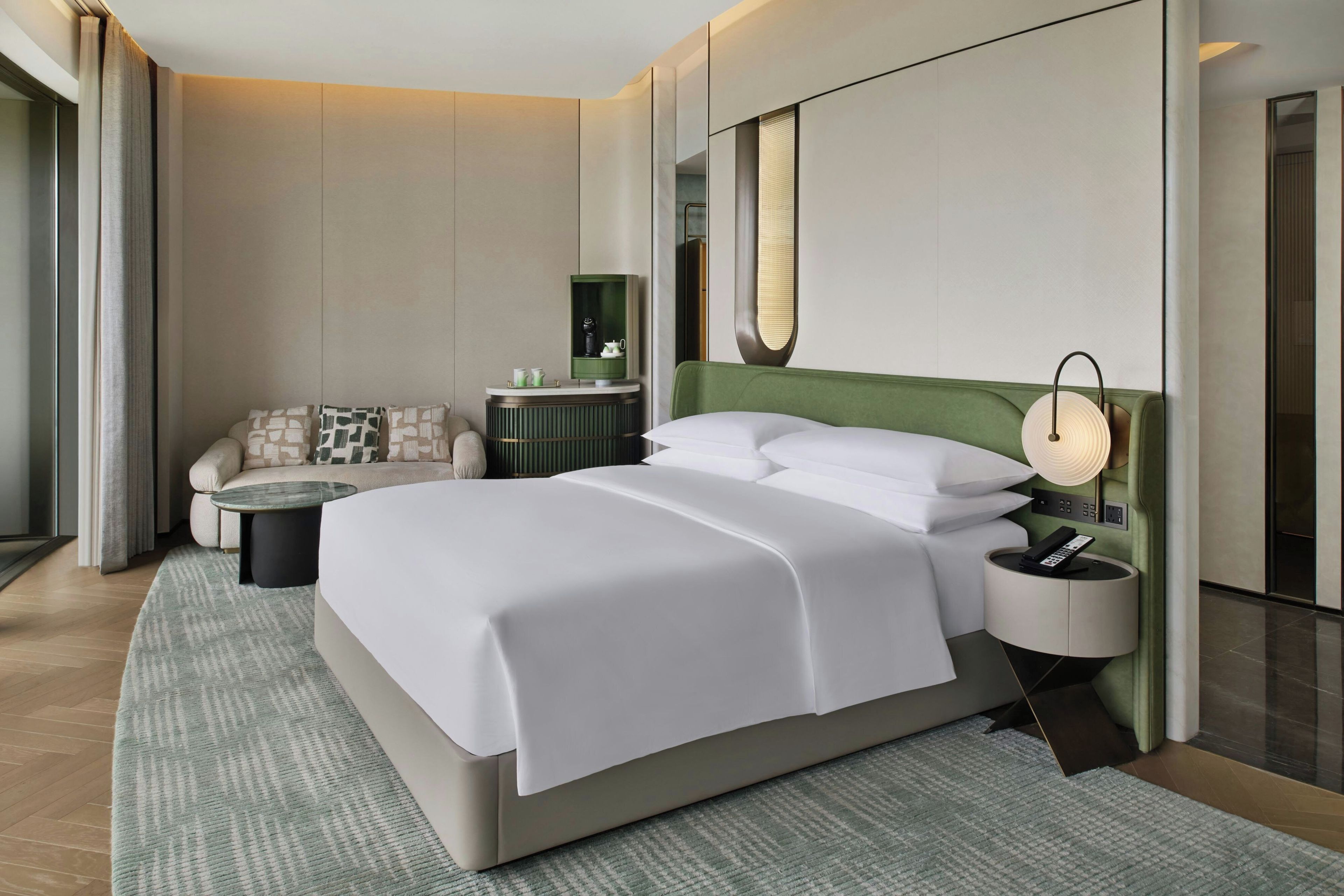 A modern hotel room with a large bed, a sofa, a side table, a nightstand, and a mirror.