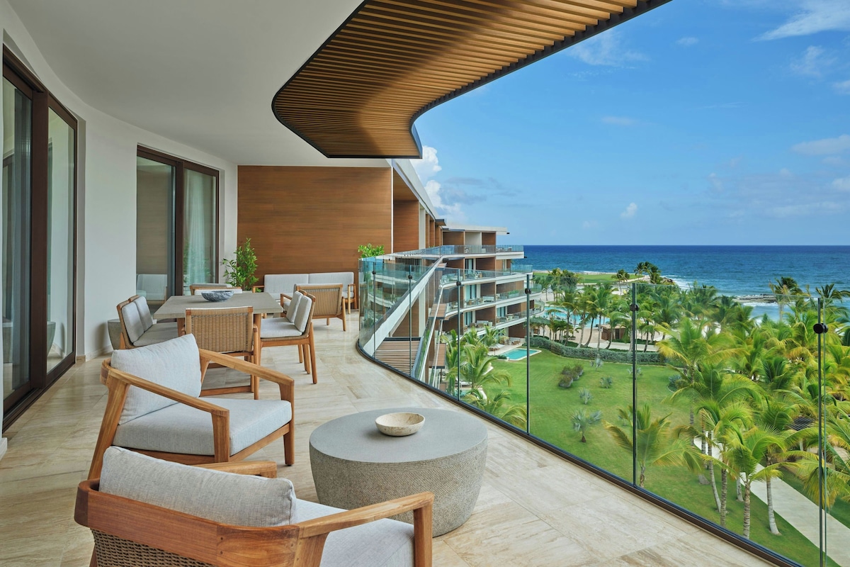 A modern outdoor balcony with a view of the ocean, furnished with wooden chairs and a round table.
