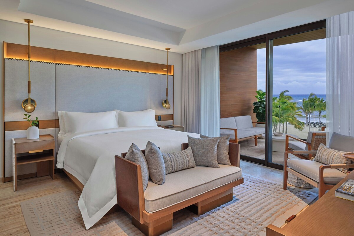 A modern hotel room with a large bed, a sofa, a desk, and a view of the ocean.