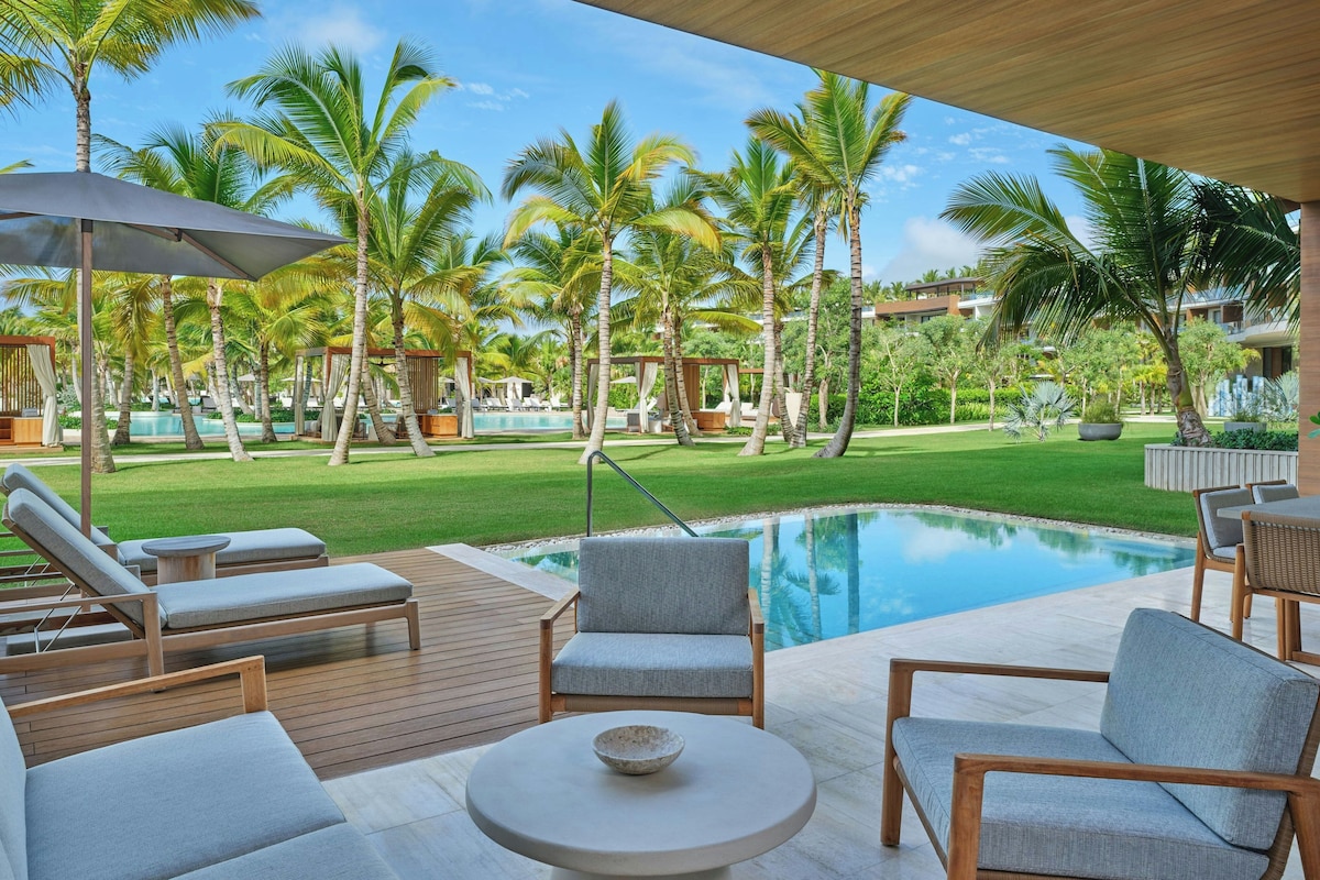 A poolside area with lounge chairs and a table, surrounded by palm trees and a clear sky.