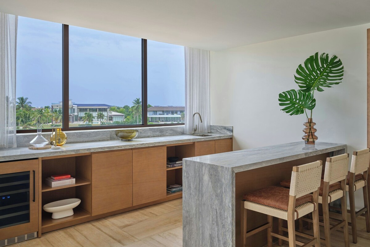 A modern kitchen with a marble countertop, wooden cabinets, a built-in sink, and a large window offering a view of palm trees and buildings.
