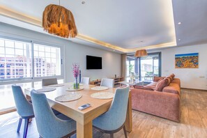 A modern living room with a sofa, dining table, chairs, and a TV mounted on the wall. - Mohammed VI | Elegant 3-bedroom apartment with a terrace (Maroc)