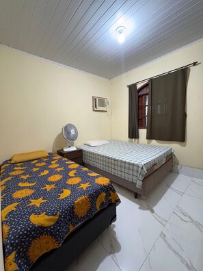 A bedroom with two beds, a standing fan, an air conditioning unit, and a window with curtains. - Beautiful house near the beach. (Cabo Frio)