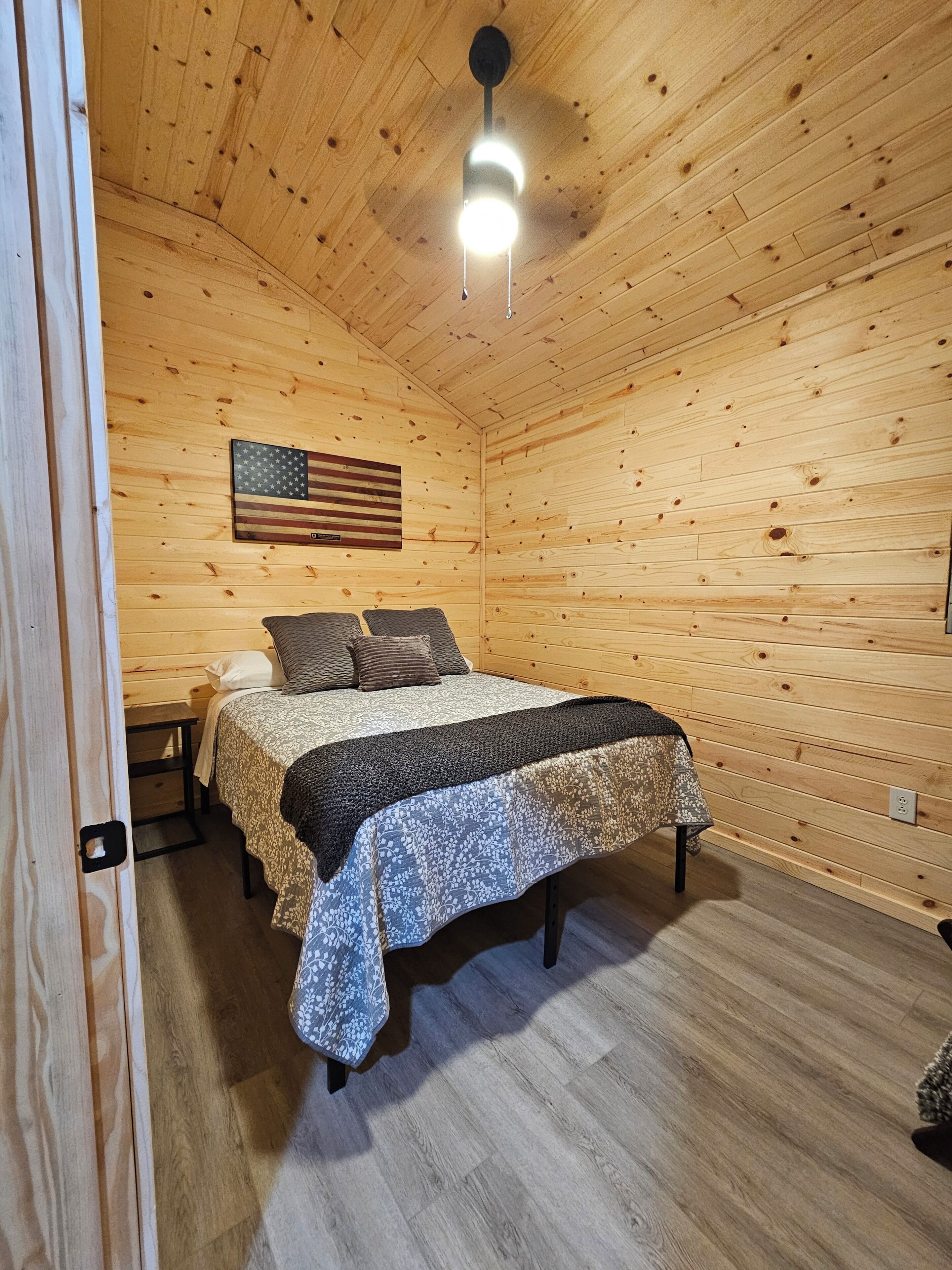 Une pièce aux murs lambrissés, avec un lit, un ventilateur de plafond et un drapeau accroché au mur.