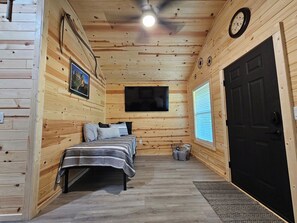 Un dormitorio de cabaña de madera con una cama, un ventilador de techo, un televisor montado y una puerta negra.