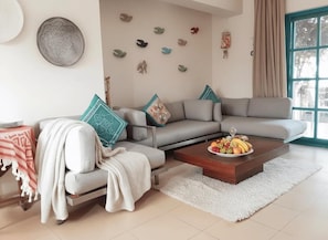 A living room with a sectional sofa, a coffee table with fruit, a patterned throw, and wall decorations.