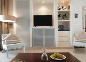 A living room with a flat-screen TV, two white armchairs, a coffee table with a glass decanter and fruit, and a bookshelf with books and decorative items.