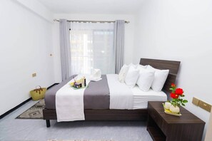 Ein ordentlich bezogenes Bett mit weißer Bettwäsche, ein Nachttisch mit Blumen in einer Vase und ein Fenster mit transparenten Vorhängen.